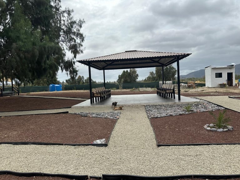 Modern covered picnic area with bench seating under a black pergola, surrounded by gravel pathways and landscaping on a rural property